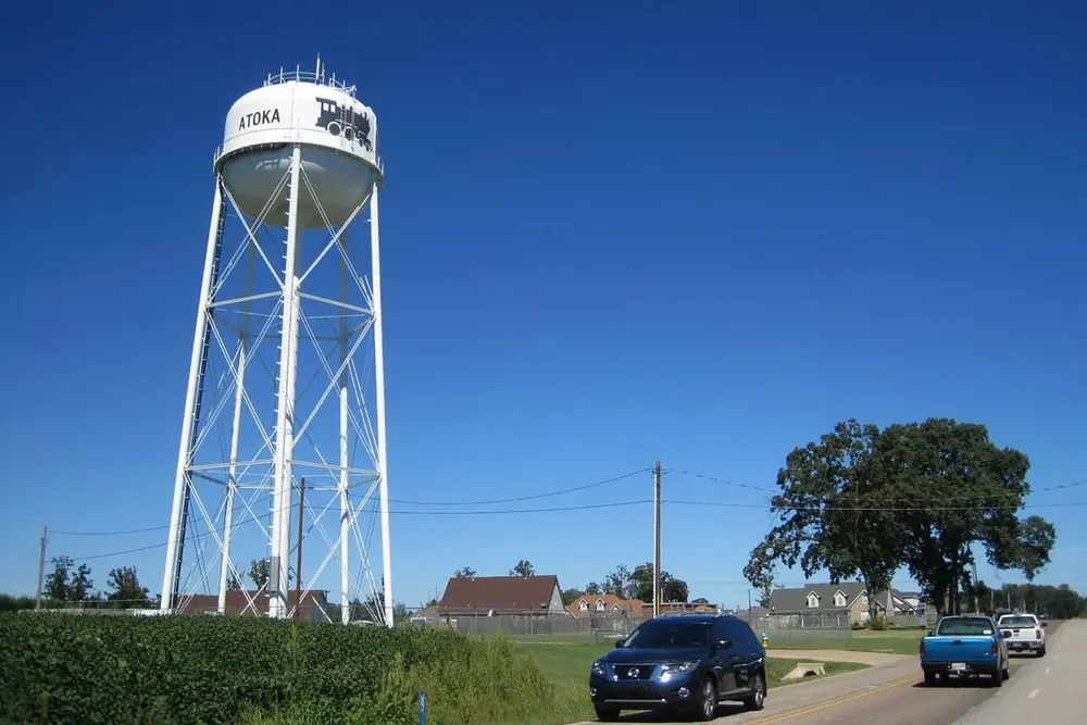 White water tower with an Atoka label rising above a field and a two-lane road in Atoka, Tennessee